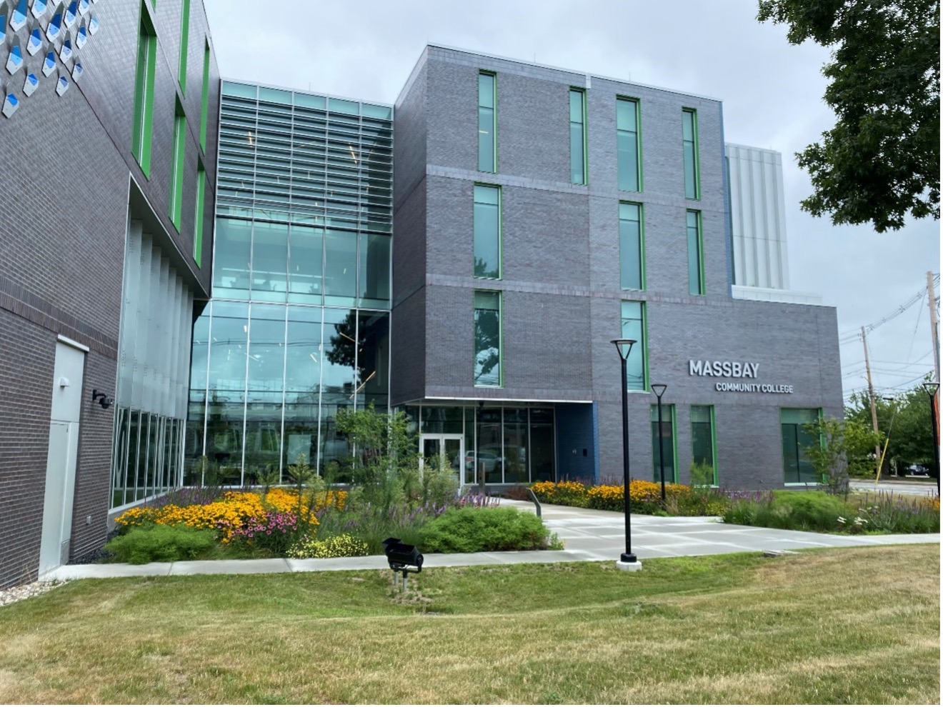 Modern MassBay Community College building with gray brick facade, green-trimmed vertical windows, a glass entryway, and landscaped grounds featuring walkways, grass, and yellow and purple flowers.